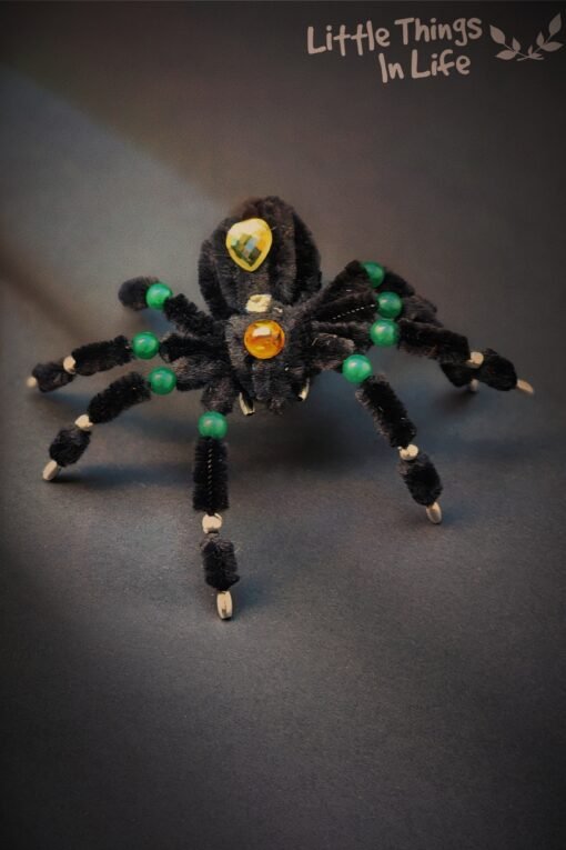 Handmade black spider decoration made with pipe cleaners, featuring crystal bead embellishments in green, amber, and yellow. Perfect for Halloween decor or a unique desk ornament.